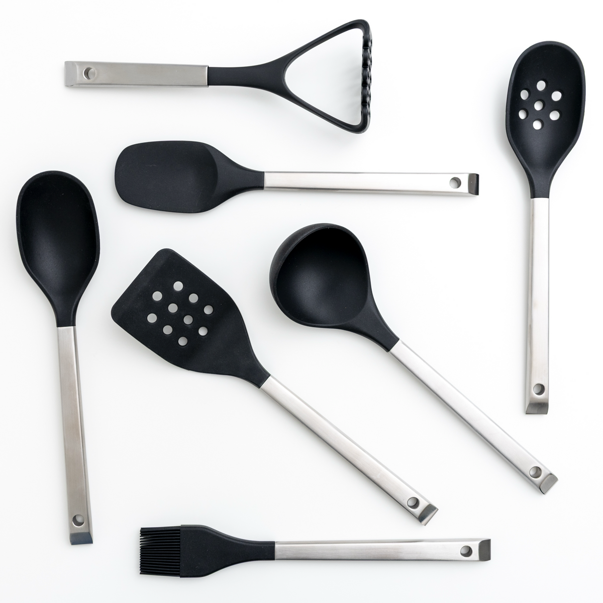 Seven kitchen utensils with black silicone heads and silver metal handles, including a potato masher, spoons, spatulas, and a brush, arranged on a white background.
